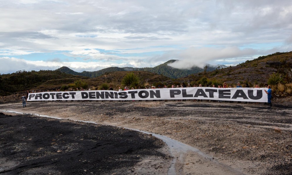A line of people holding a white banner with words in black "Save Denniston Plateau"