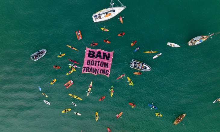 A bird's eye view of kayaks and boats on the water surrounding a giant bright pink banner with the words 'Ban Bottom Trawling'