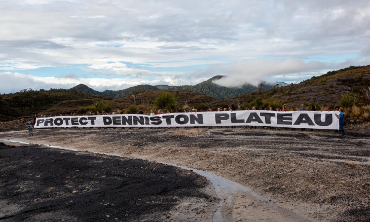 A line of people holding a white banner with words in black "Save Denniston Plateau"